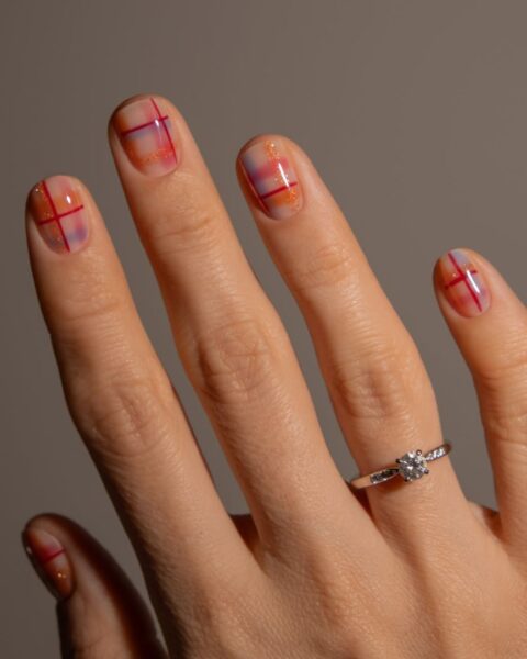 Delikatny i nowoczesny manicure w kratkę na krótkich paznokciach o zaokrąglonym kształcie. Wzór kraty jest minimalistyczny, stworzony z cienkich, fuksjowych i pomarańczowych linii na przeźroczystej lub bardzo jasnej beżowej bazie, z subtelnym, brokatowym akcentem. Na palcu serdecznym widoczny jest srebrny pierścionek zaręczynowy z centralnym, okrągłym diamentem i mniejszymi kryształkami na obrączce. Jest to stylowa i dyskretna inspiracja na świąteczny nail art.