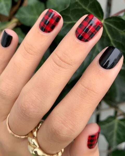Efektowny i wyrazisty manicure w kratkę (buffalo check) w stylu rockowym lub jesienno-zimowym, na krótkich, kwadratowych paznokciach. Dwa paznokcie (wskazujący i serdeczny) są pokryte gęstą, czerwoną i czarną kratą, a pozostałe trzy są pomalowane na jednolity, lśniący, głęboki czarny kolor. Dłoń ozdobiona jest masywnymi, złotymi pierścionkami w nowoczesnym stylu, a tłem dla zdjęcia jest soczyście zielony bluszcz.