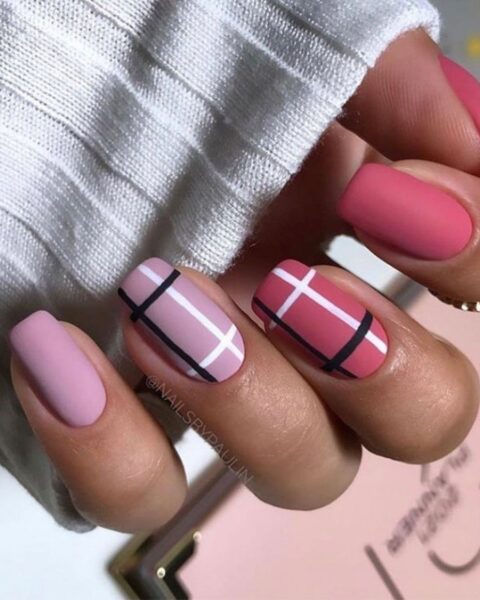 Uroczy i nowoczesny manicure w kratkę na krótkich, zaokrąglonych paznokciach, utrzymany w odcieniach matowego różu i fuksji. Paznokcie mały i serdeczny są jednolicie różowe, a środkowy i wskazujący mają wzór kraty z grubych, czarnych i białych linii na jaśniejszej, różowej bazie. Stylizacja ma matowe wykończenie i idealnie pasuje do białego, prążkowanego swetra. Inspiracja modnym nail art na jesień/zimę.