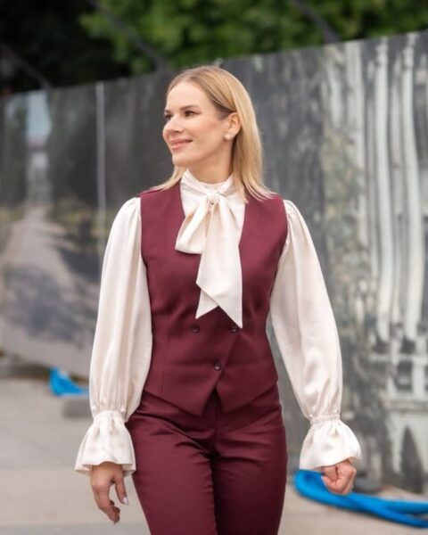 Marta Nawrocka w szykownym, retro zestawie: bordowym garniturze (spodnie i kamizelka dwurzędowa) oraz jedwabnej, kremowej bluzce z żabotem/kokardą. Uśmiechnięta idzie ulicą, na tle czarno-białego baneru przedstawiającego architekturę. Elegancki i klasyczny styl z nutą vintage.