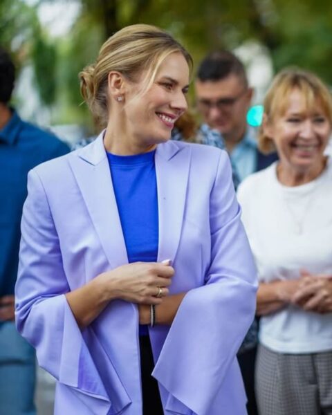 Marta Nawrocka uśmiecha się, patrząc w bok, idąc ulicą w tłumie. Ma na sobie modną, lawendową (liliową) marynarkę oraz chabrowy (kobaltowy) top, który kontrastuje z jej jasną marynarką. Kolorowy i wyrazisty look, idealny do stworzenia inspirującego artykułu o modzie. W tle widać uśmiechniętą kobietę w białej koszulce i inne osoby.