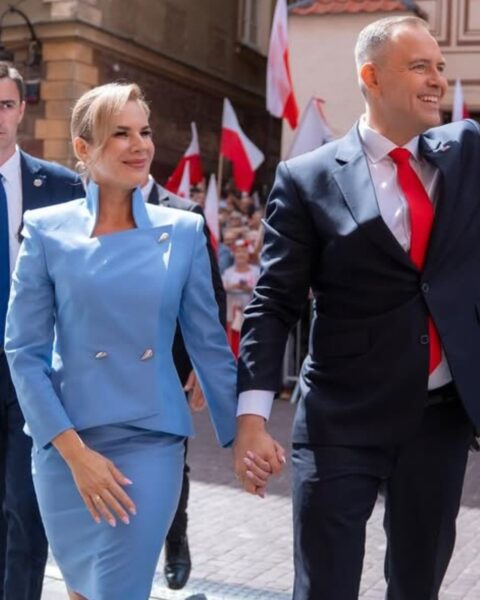 Marta Nawrocka w szykownym, błękitnym garsonce ze spódnicą ołówkową i żakietem z podniesionym kołnierzem i srebrnymi, dwurzędowymi guzikami, trzyma za rękę męża (lub konkretną postać polityczną) w granatowym garniturze i czerwonym krawacie. Para uśmiecha się, maszerując w tłumie z polskimi flagami w tle. Elegancki i klasyczny styl.