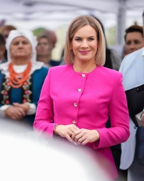 Marta Nawrocka w intensywnie różowej, eleganckiej marynarce ze złotymi guzikami, uśmiechnięta, stoi w tłumie podczas dożynek lub wydarzenia folklorystycznego. W tle widać osoby w tradycyjnych strojach ludowych (np. chusta, korale) oraz namioty. Wyrazisty, kobiecy styl.