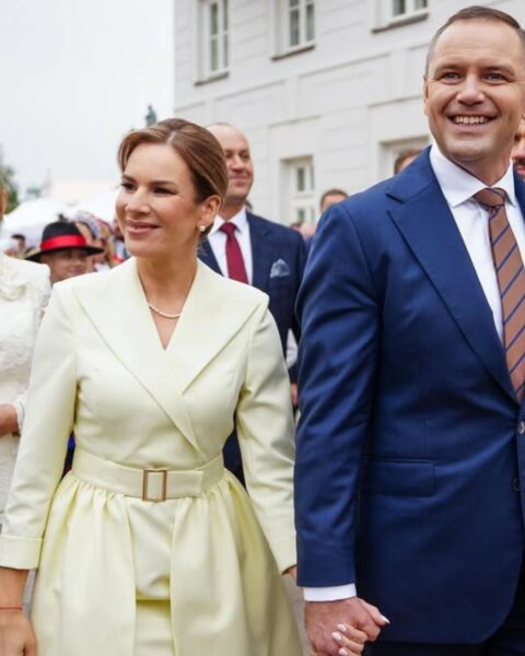 Marta Nawrocka w eleganckiej, kremowej (lub bardzo jasnej żółtej) sukience z dekoltem w serek i szerokim paskiem w talii, trzyma za rękę męża (lub konkretną postać polityczną) w granatowym garniturze i brązowym krawacie. Para uśmiecha się, idąc wśród tłumu na Dożynkach Prezydenckich w Warszawie. W tle widać budynek w klasycznym stylu oraz kobiety w tradycyjnych, ludowych strojach. Subtelna elegancja w stylu pierwszej damy.