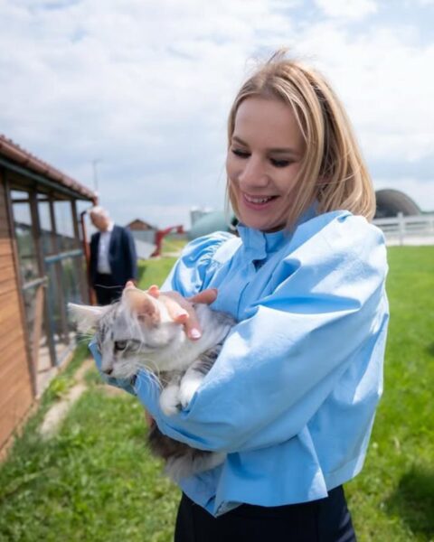 Marta Nawrocka, uśmiechnięta, trzyma w ramionach dużego, puszystego kota rasy Maine Coon na tle stadniny koni arabskich Klikowa Arabians. Ma na sobie modną, błękitną koszulę z bufiastymi rękawami oraz ciemne spodnie. Całość stylizacji w duchu elegancji casual. W tle widać budynki stajni i kobietę w szarym garniturze. Radosny portret, bliskość ze zwierzętami.