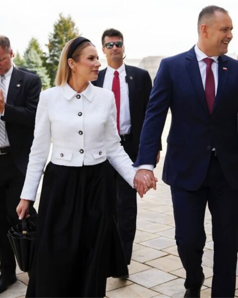 Marta Nawrocka w eleganckim, dwukolorowym zestawie – białej marynarce/żakiecie z kołnierzykiem i czarnej, długiej spódnicy – trzyma za rękę męża w granatowym garniturze i bordowym krawacie. Para wychodzi z budynku w towarzystwie ochrony i zakonnika w białym habicie. Stylizacja pierwszej damy to połączenie klasyki i mody. Sprawdź, marki które nosi.
