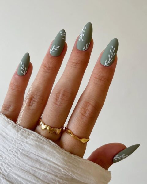 Zbliżenie na dłoń z długimi, migdałowymi paznokciami w pięknym, szałwiowym odcieniu zieleni (pastelowa, zgaszona zieleń). Manicure ma matowe wykończenie, a na każdej płytce widoczny jest delikatny, biały wzór liści lub gałązki. Dłoń zdobią dwa cienkie, złote pierścionki, jeden w kształcie serc. Fragment białego, luźnego materiału (rękaw koszuli) zakrywa część dłoni. Tło jest jasne i neutralne.