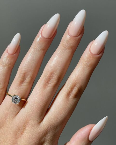 Zbliżenie na dłoń z długimi, szpiczastymi paznokciami, wykonanymi w technice French Ombre (delikatne przejście bieli w jasnoróżową bazę). Manicure jest wykończony na wysoki połysk. Dłoń ozdabia złoty pierścionek zaręczynowy z dużym, błyszczącym diamentem. Tło jest gładkie i szare.