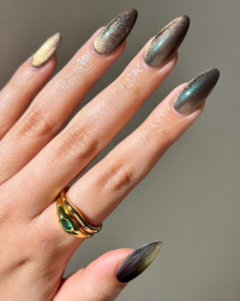 Zbliżenie na dłoń z długimi, owalnymi/migdałowymi paznokciami, na których wykonano modny manicure z efektem Cat Eye (paznokcie magnetyczne). Kolor lakieru zmienia się pod wpływem światła, od ciemnego brązu/grafitu po opalizujący, złoto-zielony poblask. Jeden paznokieć (na kciuku) ma intensywnie złoty blask. Dłoń zdobi masywny, złoty pierścionek w kształcie węża ze szmaragdowym oczkiem.