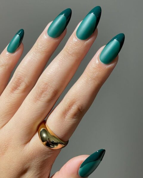 Zbliżenie na dłoń z długimi, migdałowymi paznokciami, na których wykonano geometryczny French Manicure w odcieniach szmaragdu. Większość paznokci jest pokryta matowym, butelkowym/szmaragdowym lakierem. Końcówka (tip) paznokcia, oddzielona od reszty łukiem, jest pomalowana na ten sam kolor, ale wykończona na wysoki połysk. Dłoń zdobi masywny, polerowany, złoty pierścionek o opływowym kształcie. Tło jest neutralne, szare.