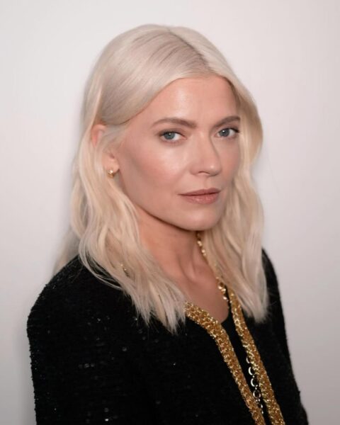 Opis alt do tego zdjęcia:Zbliżenie na Kate Young w czarnej, cekinowej kurtce Chanel ze złotym łańcuchem. Ma platynowy blond włosy ułożone w delikatne fale z przedziałkiem na środku, subtelny makijaż, który podkreśla jej niebieskie oczy oraz mały, złoty kolczyk w uchu. Elegancka stylizacja i modna koloryzacja są świetną inspiracją dla kobiet z klasą.