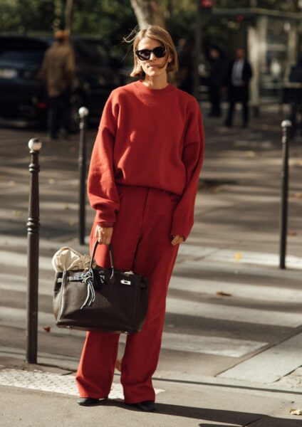 Kobieta w jesiennym, monochromatycznym zestawie street style. Stylizacja składa się z obszernego, dzianinowego swetra i szerokich spodni w intensywnym kolorze terakoty lub spalonej czerwieni. Uzupełnieniem looku jest luksusowa, czarna torebka typu Birkin i eleganckie, czarne okulary przeciwsłoneczne. Inspiracja na komfortowy i stylowy strój na chłodne dni.