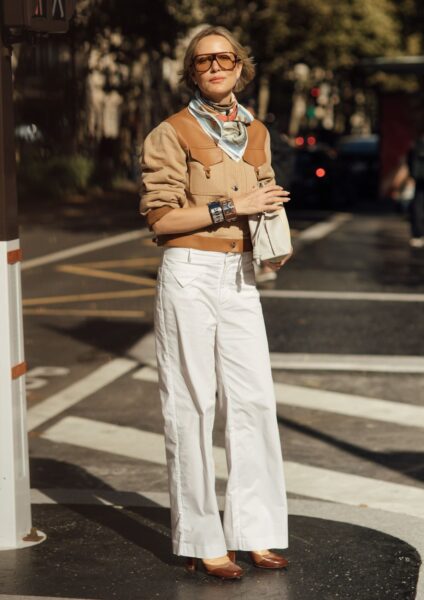 Kobieta w stylowym zestawie inspirowanym modą retro. Szerokie, białe spodnie z wysokim stanem zestawione z brązową kurtką typu bomber lub ramoneska z beżowymi akcentami. Szyku dodaje jedwabna apaszka zawiązana pod szyją, masywne bransolety i stylowe okulary przeciwsłoneczne z brązowymi szkłami. Całość uzupełniają błyszczące, brązowe czółenka i mała, biała torebka.