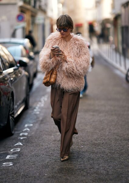 Kobieta w stylowym zestawie street style. Luksusowy, puszysty płaszcz w odcieniu pudrowego różu zestawiony z szerokimi, brązowymi spodniami z wysokim stanem. Całość uzupełnia pleciona karmelowa torebka, okulary przeciwsłoneczne i subtelne, złote czółenka. Inspiracja na szykowny i wyrazisty, ale jednocześnie elegancki jesienny look.
