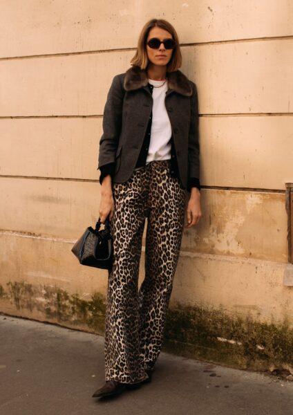 Kobieta w eleganckim, drapieżnym zestawie street style. Stylizacja opiera się na szerokich spodniach w wyrazisty motyw panterki, połączonych z klasycznym, szarym żakietem z futrzanym kołnierzem i prostym białym T-shirtem. Uzupełnieniem looku jest mała, czarna torebka, naszyjnik z pereł i okulary przeciwsłoneczne. Idealna inspiracja dla kobiet ceniących modową odwagę z klasą.