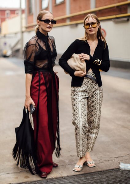 Dwie stylowe kobiety w zmysłowych zestawach street style z Mediolanu. Po lewej: czarna, transparentna koszula i długie, bordowe spodnie z frędzlami, odsłaniające elegancką czarną bieliznę. Po prawej: czarny, krótki kardigan, spodnie w wężowy wzór, puszysta beżowa torebka i okulary z żółtymi szkłami. Inspiracja odważnym, ale wyrafinowanym stylem.