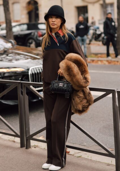 Kobieta w sportowo-eleganckim zestawie street style. Luźny, brązowy komplet dresowy z kolorowymi akcentami przy kołnierzu, połączony z modnym, czarnym kapeluszem typu bucket hat. Luksusowy akcent stanowi futrzany szal w odcieniu brązu i pikowana torebka z logo Miu Miu. Białe sneakersy dopełniają wygodny i szykowny, jesienny look.