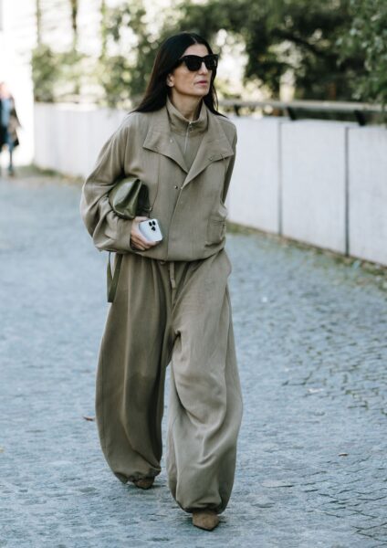 Kobieta w beżowym, monochromatycznym kombinezonie o luźnym, oversize'owym kroju. Obszerny zestaw, przypominający modny dres lub cargo, uszyty z lejącego materiału, stanowi idealny przykład minimalistycznego i komfortowego szyku. Stylizację uzupełnia oliwkowa torebka, czarne okulary przeciwsłoneczne i beżowe botki na obcasie. Inspiracja nowoczesną elegancją i wygodą.