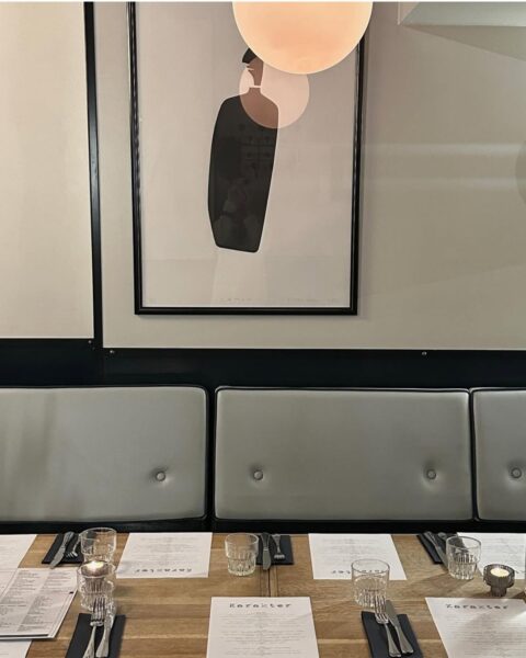Karakter – Kraków. Minimalistyczny, nowoczesny stół w restauracji, nakryty do kolacji z kartami menu Karakter i eleganckimi kieliszkami, z rzędem tapicerowanych sof w jasnoszarym kolorze na tle ściany, nad którymi wisi awangardowy plakat w czarnej ramie oraz designerska lampa o kulistym kształcie.