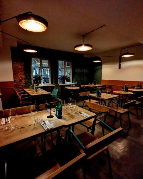 AleWino – Warszawa. Kameralne wnętrze restauracji w wieczornym półmroku, z industrialnymi lampami emitującymi ciepłe, pomarańczowe światło, rzędy drewnianych stolików nakrytych do kolacji, z widoczną ceglaną ścianą i ciemnozielonymi akcentami – stylowa, przytulna przestrzeń na wieczorne spotkanie przy winie.