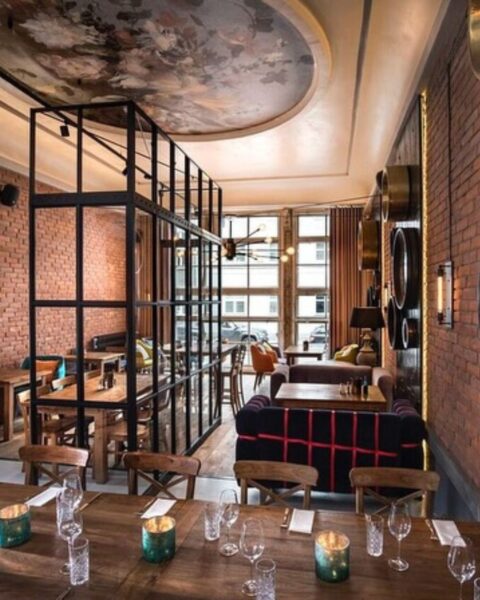 Canis – Gdańsk. Eleganckie wnętrze restauracji w stylu industrialnym z elementami vintage, z ceglanymi ścianami i panelami z ciemnego drewna, na których wiszą okrągłe lustra w metalowych ramach, pośrodku industrialne przeszklenie, a nad sufitem ozdobny plafon z malowidłem – stoły nakryte do kolacji w ciepłej, intymnej atmosferze.