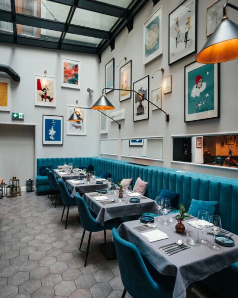 Filipa 18 Food Wine Art – Kraków. Nowoczesne wnętrze restauracji ze szklanym dachem i galerią obrazów na jasnej ścianie, turkusowe welurowe sofy i krzesła przy stolikach nakrytych szarymi obrusami, tworzące elegancką i artystyczną atmosferę, idealną na spotkanie przy wykwintnym posiłku.