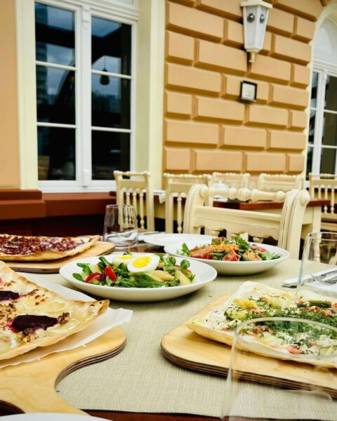 Starka – Opole. Stolik na tarasie restauracji z widokiem na elegancką żółtą fasadę budynku, na którym stoją talerze z różnymi rodzajami tart flambirowanych (flammkuchen), w tym wersje z burakiem i łososiem oraz świeże sałatki z jajkiem i warzywami, serwowane w słoneczne popołudnie – idealne miejsce na stylowy lunch.