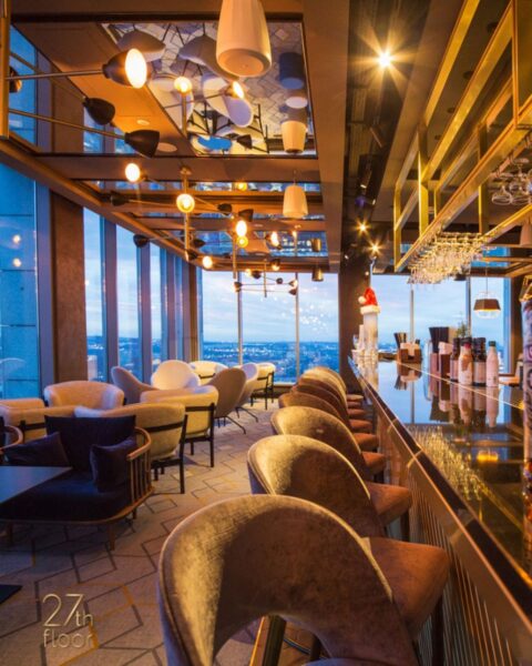 27th Floor – Katowice. Luksusowy bar na 27. piętrze z panoramicznym widokiem na miasto o zachodzie słońca, lśniący złoty blat barowy i złote regały, rząd welurowych hokerów w kolorze beżu, stoliki w strefie lounge z granatowymi fotelami i designerskimi lampami – stylowe miejsce na eleganckiego drinka na wysokości.