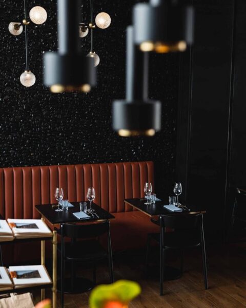 Campo Modern Grill – Wrocław. Nowoczesne wnętrze restauracji w ciemnej kolorystyce, z pomarańczową, pikowaną sofą i czarnymi stolikami dla dwojga, na tle efektownej, trójwymiarowej czarnej ściany, oświetlone przez designerskie lampy wiszące z matowego metalu – stylowa i intymna przestrzeń na elegancką kolację.