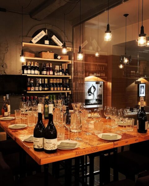 Muga Restaurant – Poznań. Przygotowany stół do degustacji wina w kameralnym, stylowym wnętrzu winiarni z ceglanymi i drewnianymi elementami, zastawiony licznymi kieliszkami i butelkami wina, w tle regały wypełnione winami oraz ciepłe, industrialne oświetlenie – idealna atmosfera na wieczór z kulturą wina i degustację Furmint.