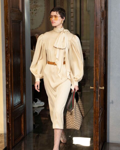 Modelka wchodząca przez ciemne, drewniane drzwi w beżowej, eleganckiej sukience midi. Sukienka ma wysoki kołnierz z dużą kokardą pod szyją oraz bufiaste rękawy. Talia jest podkreślona cienkim, brązowym paskiem. Stylizację uzupełniają cieliste sandałki na obcasie, rajstopy w subtelny wzór, duża torba z monogramem (Gucci) oraz złote, geometryczne kolczyki i okulary przeciwsłoneczne z żółtymi szkłami. Ujęcie z pokazu mody.