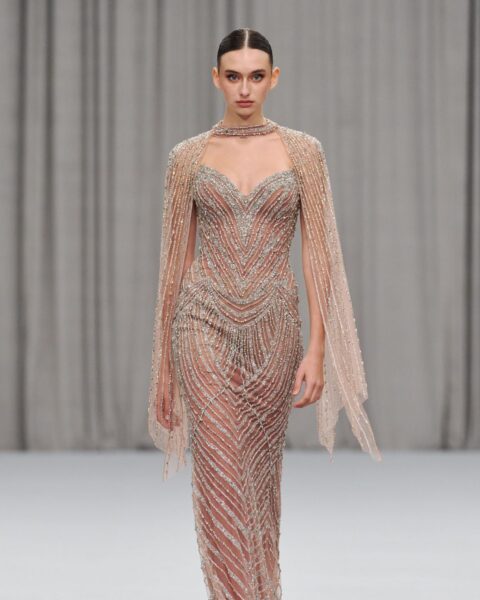 Modelka w zapierającej dech w piersiach, długiej, beżowej sukni haute couture, obficie wyszywanej błyszczącymi, srebrno-złotymi koralikami tworzącymi geometryczne, pionowe wzory. Kreacja ma gorsetowy dekolt w kształcie serca oraz dopasowaną pelerynkę/szal spływający z ramion. Modelka ma gładko zaczesane włosy i minimalistyczną biżuterię w formie chokera. Ujęcie z pokazu mody, na tle jasnoszarych zasłon.