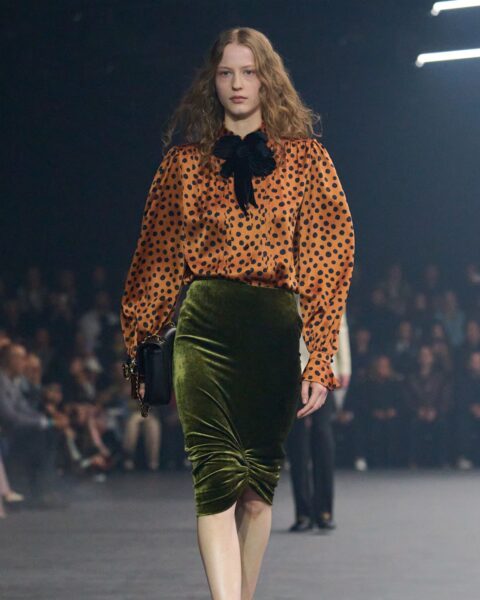 Modelka na ciemnym wybiegu w stylizacji retro: pomarańczowa bluzka w czarne kropki z bufiastymi rękawami i czarną, aksamitną kokardą pod szyją, zestawiona z obcisłą, welurową spódnicą midi w kolorze butelkowej zieleni. Spódnica jest drapowana z przodu w dolnej części. Modelka nosi czarne, klasyczne czółenka i trzyma małą, czarną torebkę na łańcuszku. Ujęcie z pokazu mody. Najważniejsze pojęcia w modzie luksusowej – słownik premium.