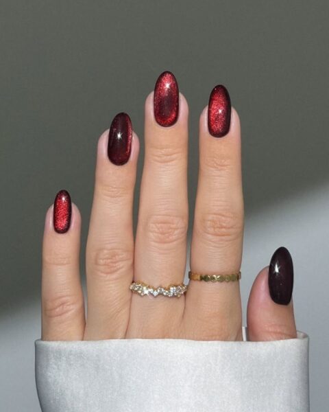 Krótkie, owalne paznokcie w kolorze głębokiej, rubinowej czerwieni lub wiśniowego wina, z intensywnym, lśniącym efektem kociego oka (cat eye). Elegancki manicure w stylu "vampire nails", idealny jako paznokcie na sezon Skorpiona 2025. Na palcach złote, geometryczne pierścionki.
