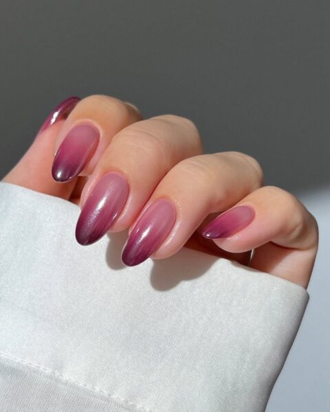 Długie, migdałowe paznokcie w stylu "lip gloss nails" z efektem ombre (gradient), przechodzącym z transparentnego, mlecznego różu przy nasadzie do głębokiego, burgundowego lub śliwkowego fioletu na końcówkach. Manicure w stylu eleganckiego, sezonowego przejścia z lata do jesieni.