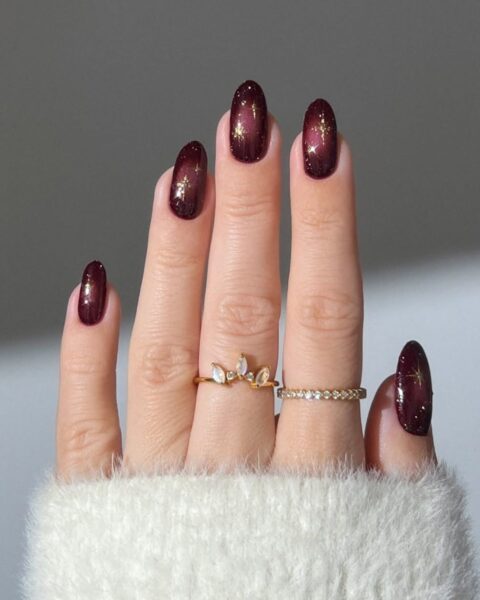Krótkie, owalne paznokcie w głębokim odcieniu śliwki lub bordo, z efektem "galaxy nails" — na ciemnej bazie widoczny jest złoty i holograficzny, rozproszony pyłek, tworzący wzór przypominający gwiezdną konstelację. Manicure idealny na eleganckie przyjęcia. Na palcach złote pierścionki.