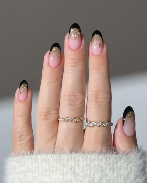Krótkie, owalne paznokcie z czarnym french manicure. Stylizacja na bazie w kolorze mlecznego różu (cover) z błyszczącym zdobieniem: gradient ze złotych, brokatowych gwiazdek, który opada z czarnej końcówki na beżową bazę. Elegancki i sylwestrowy manicure. Na palcach delikatne, biżuteryjne pierścionki.
