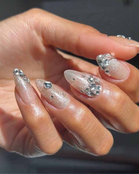 Długie, migdałowe paznokcie z bazą w kolorze mlecznego nude, pokrytą delikatnym, beżowym brokatem z efektem kociego oka lub piasku. Manicure ozdobiony bogatymi aplikacjami z dużych i małych cyrkonii i kryształków, tworzącymi efekt biżuterii 3D. Luksusowy styl "bling nails" na sezon świąteczny.