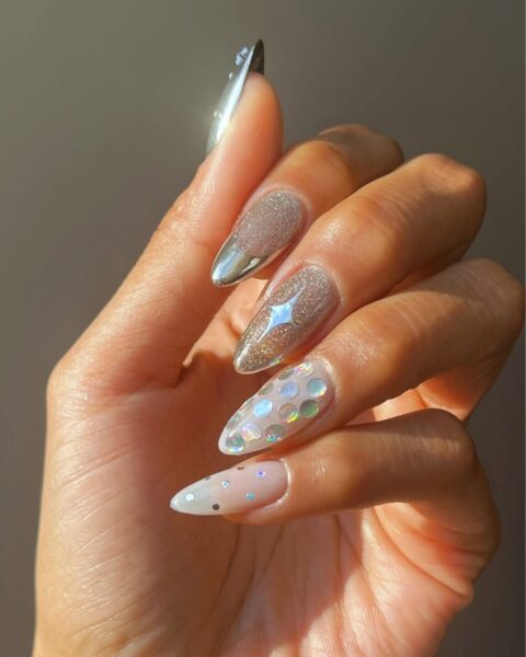 Długie, szpiczaste paznokcie z mieszanym zdobieniem w stylu "mix and match". Ozdobione brokatem w kolorze szampańskiego złota, srebrnym chromem (metaliczny french), efektem holograficznych kropek i małych, holograficznych kółek, zatopionych w mlecznej bazie. Imprezowy i błyszczący manicure na sezon świąteczny.