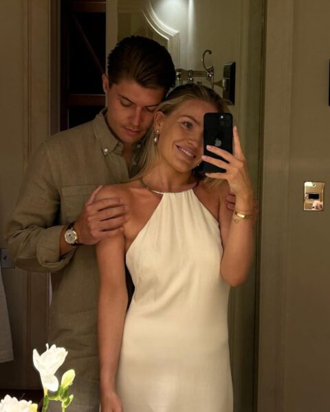 Romantyczne lustrzane selfie eleganckiej pary w ciepłym, przytulnym oświetleniu. Kobieta jest ubrana w kremową lub białą sukienkę typu halter i uśmiecha się do telefonu. Ma delikatną biżuterię – kolczyki, naszyjnik typu choker i bransoletki. Mężczyzna stoi za nią, obejmując ją ramieniem, ubrany w lnianą koszulę w kolorze beżowym lub taupe i zegarek. Oboje są elegancko ubrani, co sugeruje, że szykują się na wieczorne wyjście.