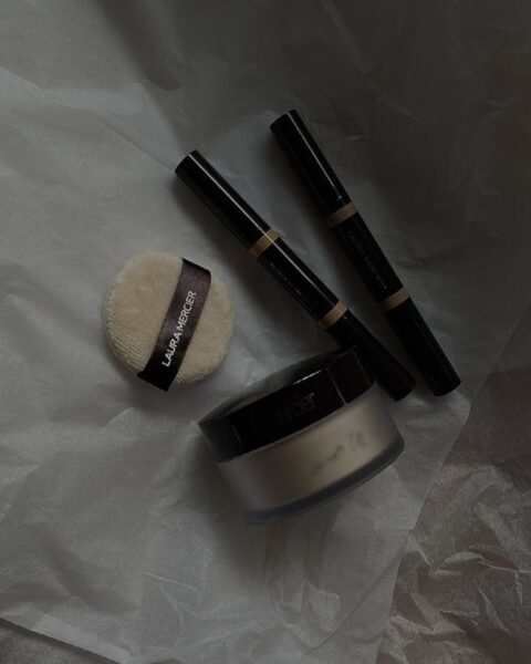 Zestaw do makijażu Laura Mercier, ułożony na pogniecionej białej bibułce: słoiczek sypkiego pudru Translucent Loose Setting Powder, aplikator typu "puszek" oraz dwa korektory w czarnych opakowaniach. Zdjęcie ma ciemny, nastrojowy filtr.