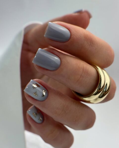Jesienny, szary manicure w jasnym, przydymionym odcieniu. Paznokcie w kształcie kwadratu z zaokrąglonymi brzegami. Dwa paznokcie są ozdobione delikatnymi płatkami złotego liścia (gold foil). Dłoń zdobi masywny, złoty pierścionek o geometrycznym kształcie.