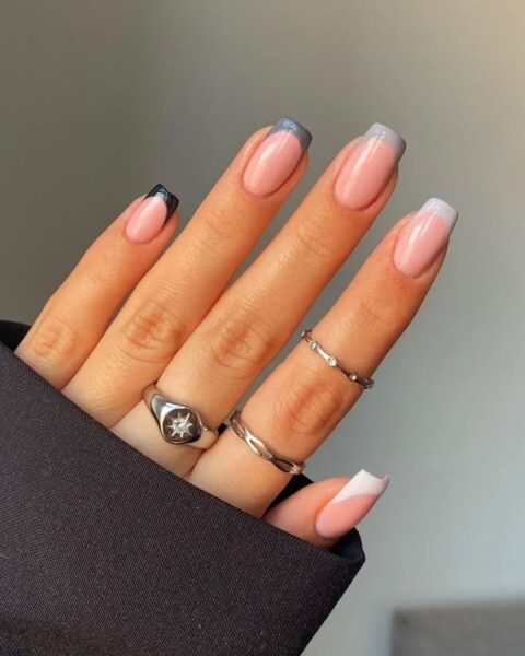 Nowoczesny french manicure z kwadratowymi paznokciami, charakteryzujący się szarym, przydymionym kolorem końcówek oraz jedną końcówką w kolorze czarnym i jedną białą, na naturalnej, mlecznoróżowej bazie. Dłoń ozdobiona srebrnymi sygnetami z cyrkoniami, w eleganckiej, czarnej marynarce, na tle rozmytego, jasnoszarego tła.