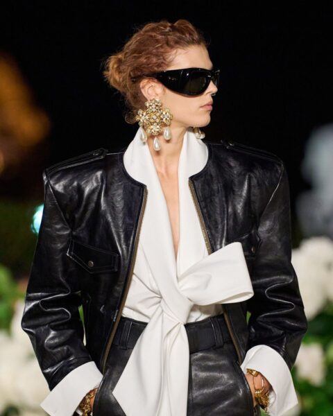 Modelka na wybiegu Saint Laurent w wieczorowej, eleganckiej stylizacji. Ma na sobie czarną, transparentną bluzkę z długimi rękawami, ciemne, szerokie spodnie z wysokim stanem oraz szeroki pasek z dużą, złotą klamrą. Uzupełnieniem looku są duże, awiatorskie okulary przeciwsłoneczne i masywne, złote kolczyki.