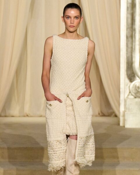 Modelka na wybiegu Chanel Haute Couture w kremowej, tweedowej mini-sukience bez rękawów z prostym dekoltem, ozdobionej złotymi guzikami i kieszeniami. Strój dopełniają beżowe, zamszowe kozaki sięgające za kolano. Cała stylizacja utrzymana jest w jasnej, eleganckiej, monokolorystycznej palecie beży i écru.