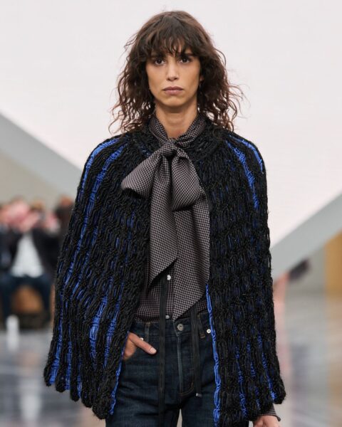 Modelka na wybiegu Dior w eleganckiej, warstwowej stylizacji. Ma na sobie czarną, ażurową pelerynę z niebieskimi akcentami, koszulę z zawiązaną pod szyją kokardą w drobną kratkę oraz ciemne jeansy z ozdobnymi sznurkami. Całość dopełnia brązowa torebka-kuferek, trzymana w dłoni.