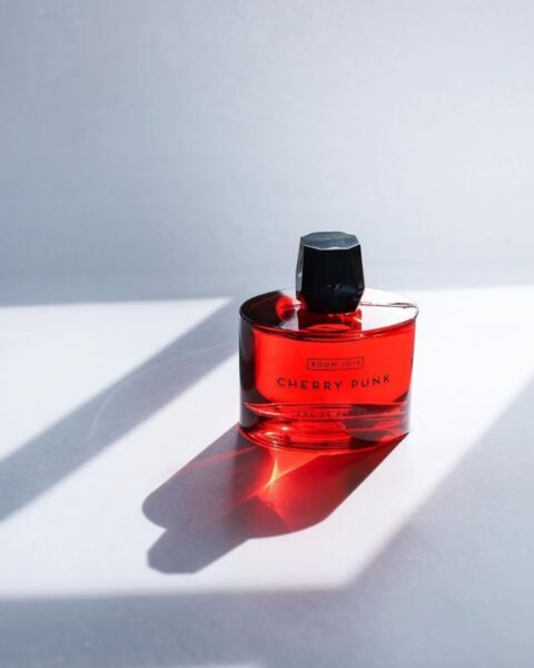 Flakon perfum Room 1015 Cherry Punk Eau de Parfum w intensywnie czerwonym kolorze stoi w mocnym, słonecznym świetle, rzucając długie cienie na jasne tło. To propozycja wiśniowych perfum na jesień 2025 o rockowym, skórzanym charakterze. Perfumy wiśniowe na jesień.
