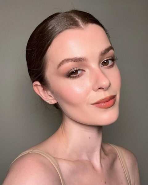 Portret kobiety w cielistym topie z włosami gładko zaczesanymi w niski kok i eleganckim, naturalnym makijażem w odcieniach nude. Brązowe cienie na powiece, rozświetlona cera i usta w kolorze brzoskwiniowego błyszczyka. Opis podkreśla efekt subtelnie podkreślonych oczu i rzęs uzyskany dzięki zastosowaniu najlepszego kosmetyku, jakim są wydłużające tusze rozdzielające rzęsy.