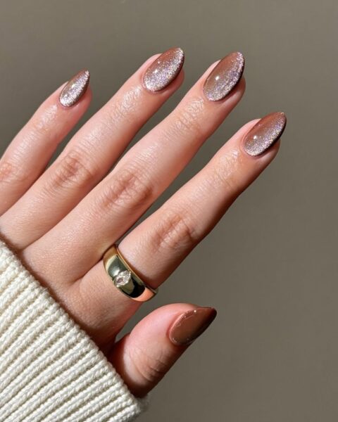 Manicure na jesień w modnym odcieniu ciepłego brązu kawowego/cappuccino. Paznokcie w kształcie owalu/migdała z lśniącym, magnetycznym (Cat Eye) brokatowym akcentem w odcieniach różowego złota lub szampana, tworzącym efekt gradientu/cienia. Dłoń otulona jasnym, kremowym swetrem z dzianiny. Na palcu serdecznym widoczny szeroki, złoty pierścionek z przezroczystym oczkiem.