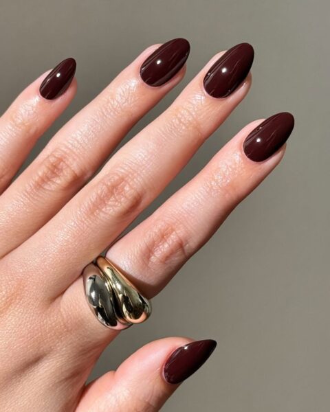 Manicure idealny na jesień i zimę. Paznokcie w kształcie migdała/owalu pokryte intensywnym, błyszczącym lakierem w kolorze głębokiego bordowego wina/ciemnej czekolady. Kolor jest klasyczny i bardzo elegancki. Na palcu serdecznym widoczny modny, duży pierścionek z dwoma metalowymi, wypukłymi elementami w kolorze srebrnym i złotym.